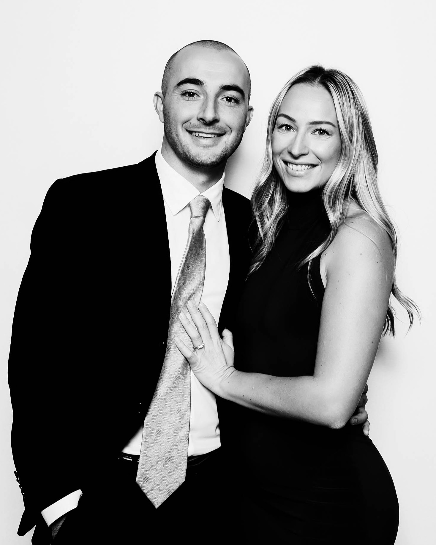 Smiling couple posing together in formal attire against a plain background, with the woman’s hand resting on the man's chest.