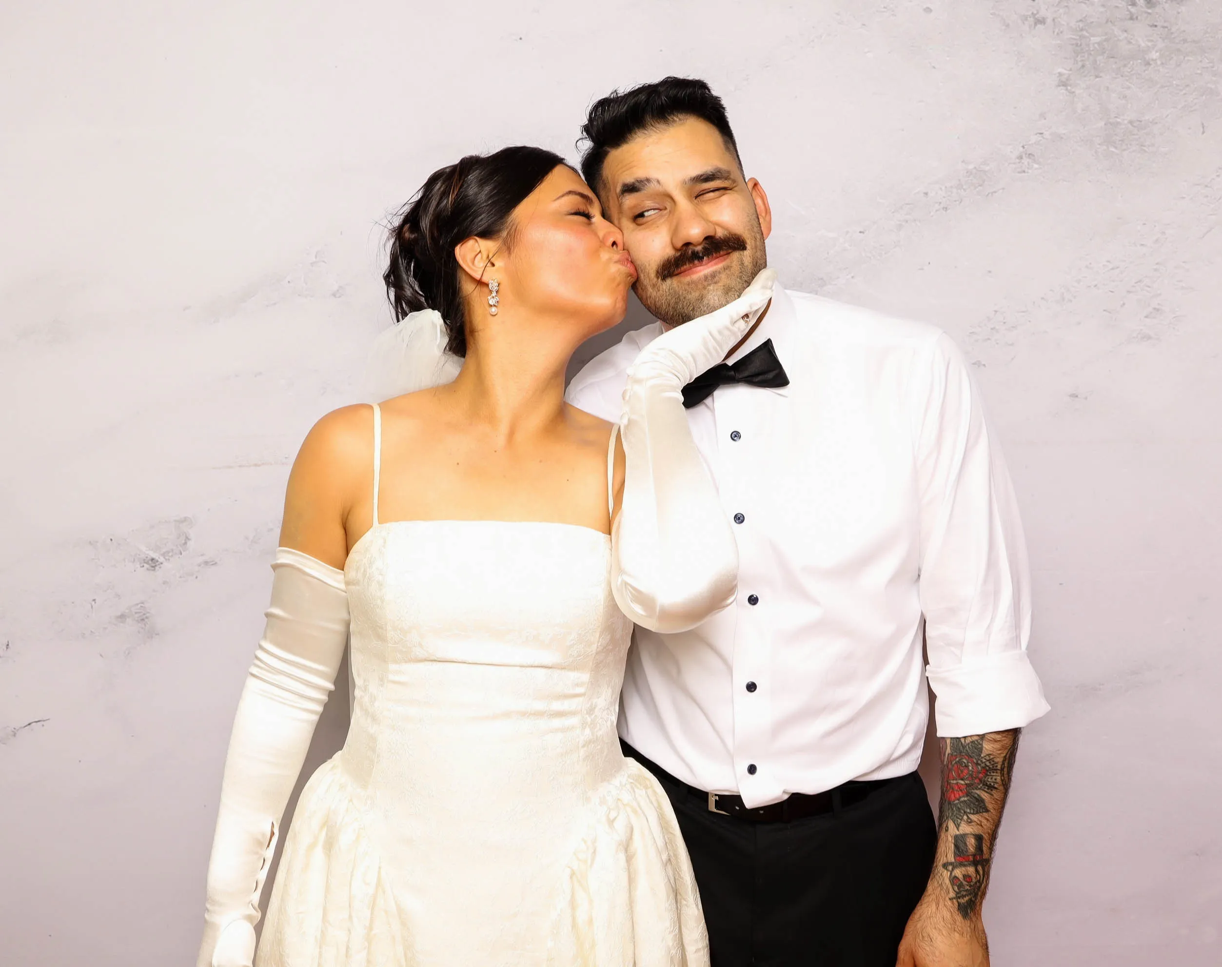 Bride in white wedding dress and gloves kissing groom with mustache and tattooed arm in a white shirt and black bow tie.