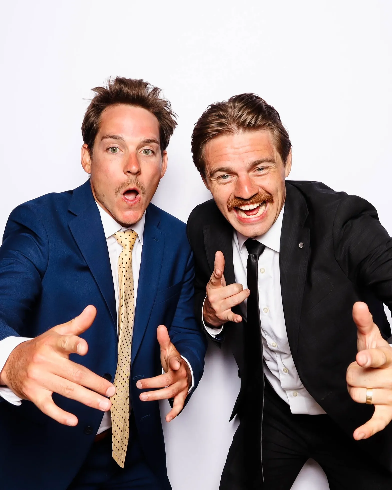 Two men in suits posing playfully with finger guns and expressive faces against a white background.