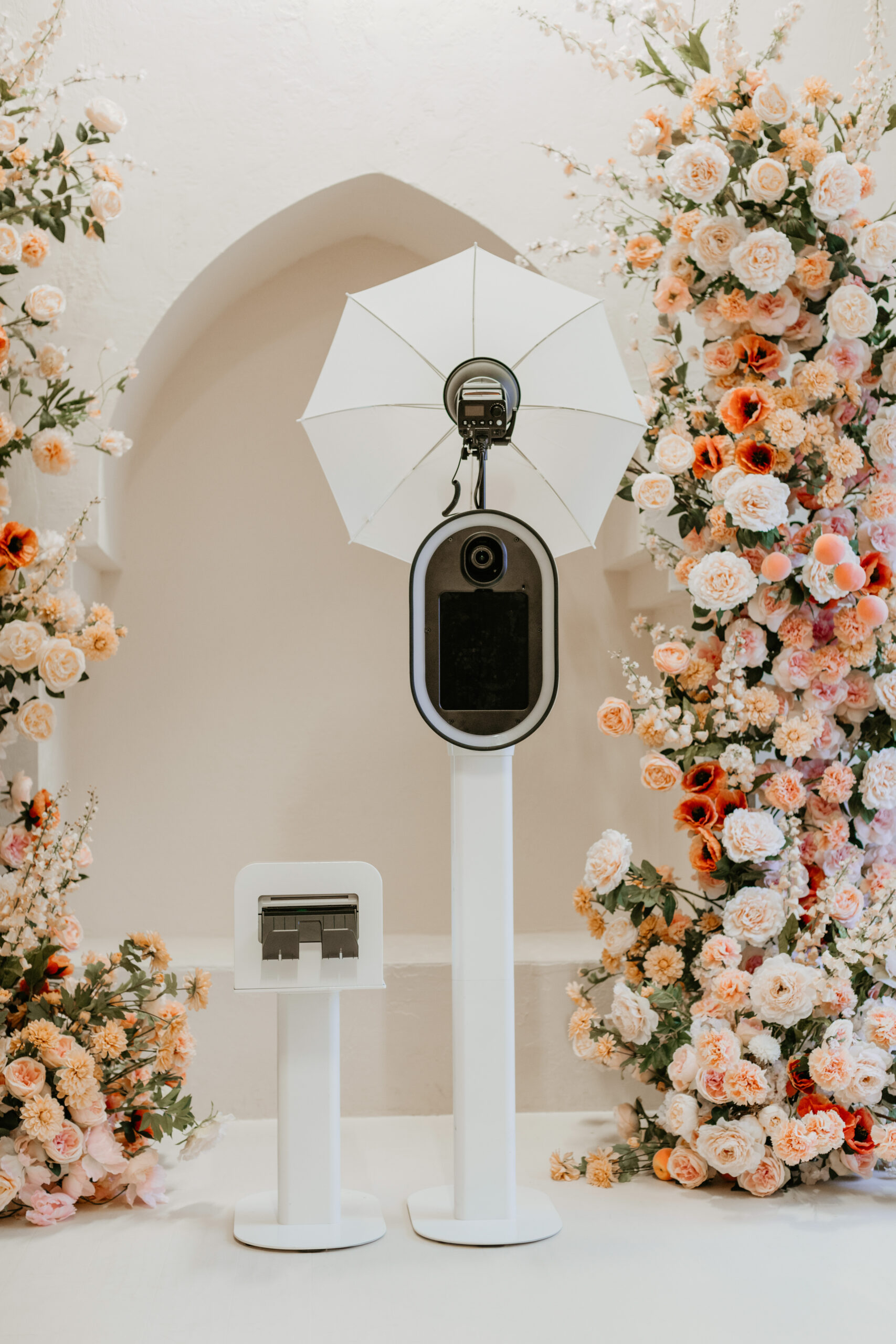 Photo booth setup with a camera on a tall white stand topped with a white umbrella, next to a small photo printer, surrounded by peach and cream floral arrangements.