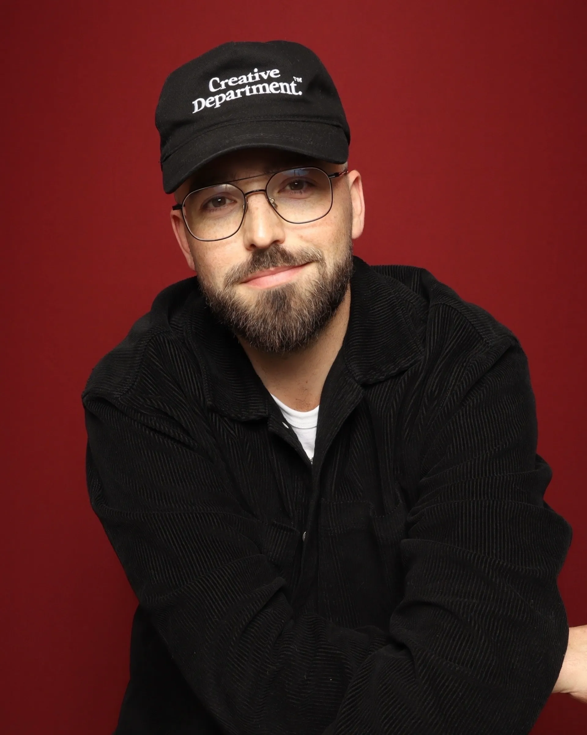 Headshot of a bearded man with a hat in front of a wine colored backdrop