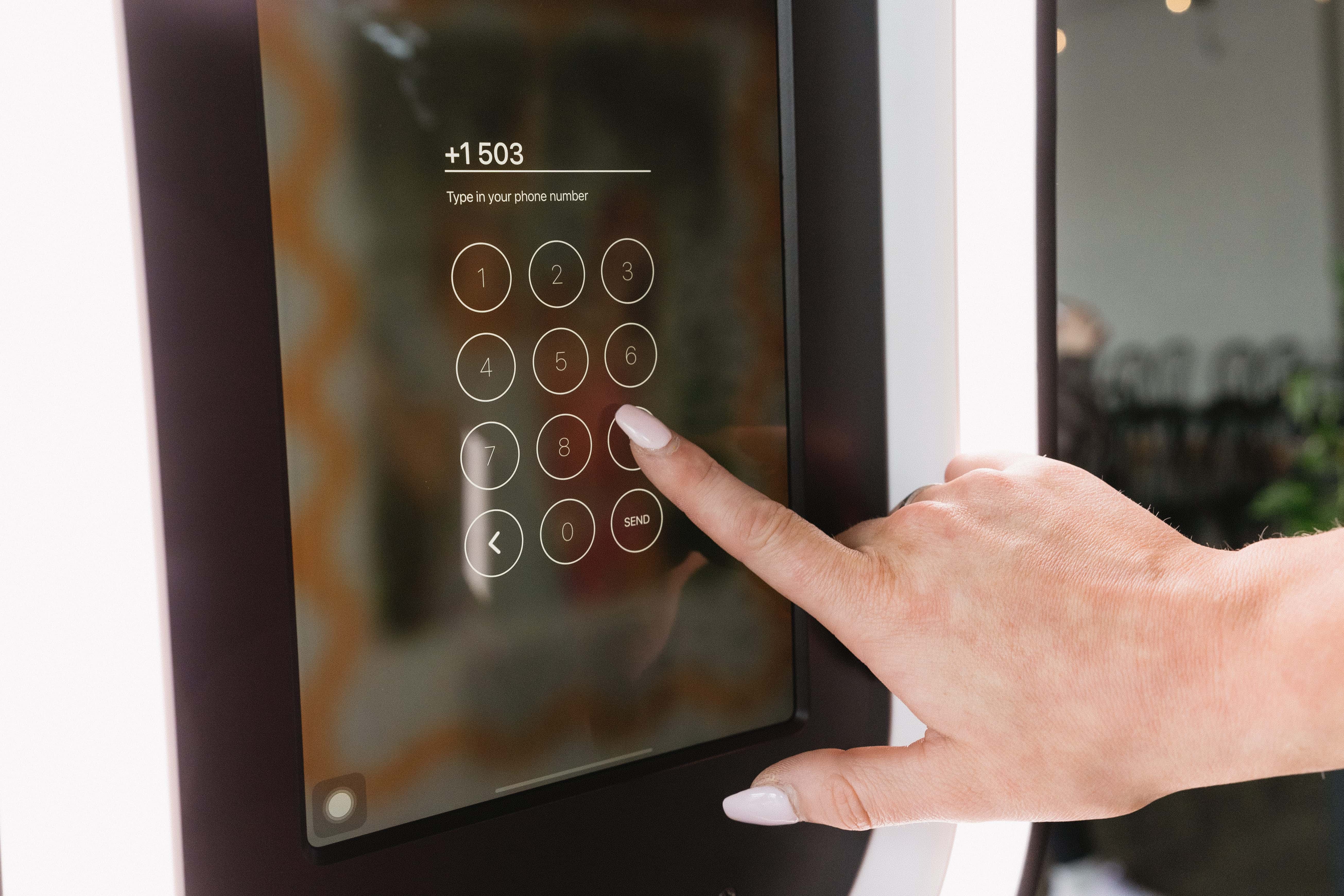 Close-up of a hand holding a smartphone displaying photo booth images of two women in winter gear next to printed photos and a drink can with a QR code sign on a clear table.