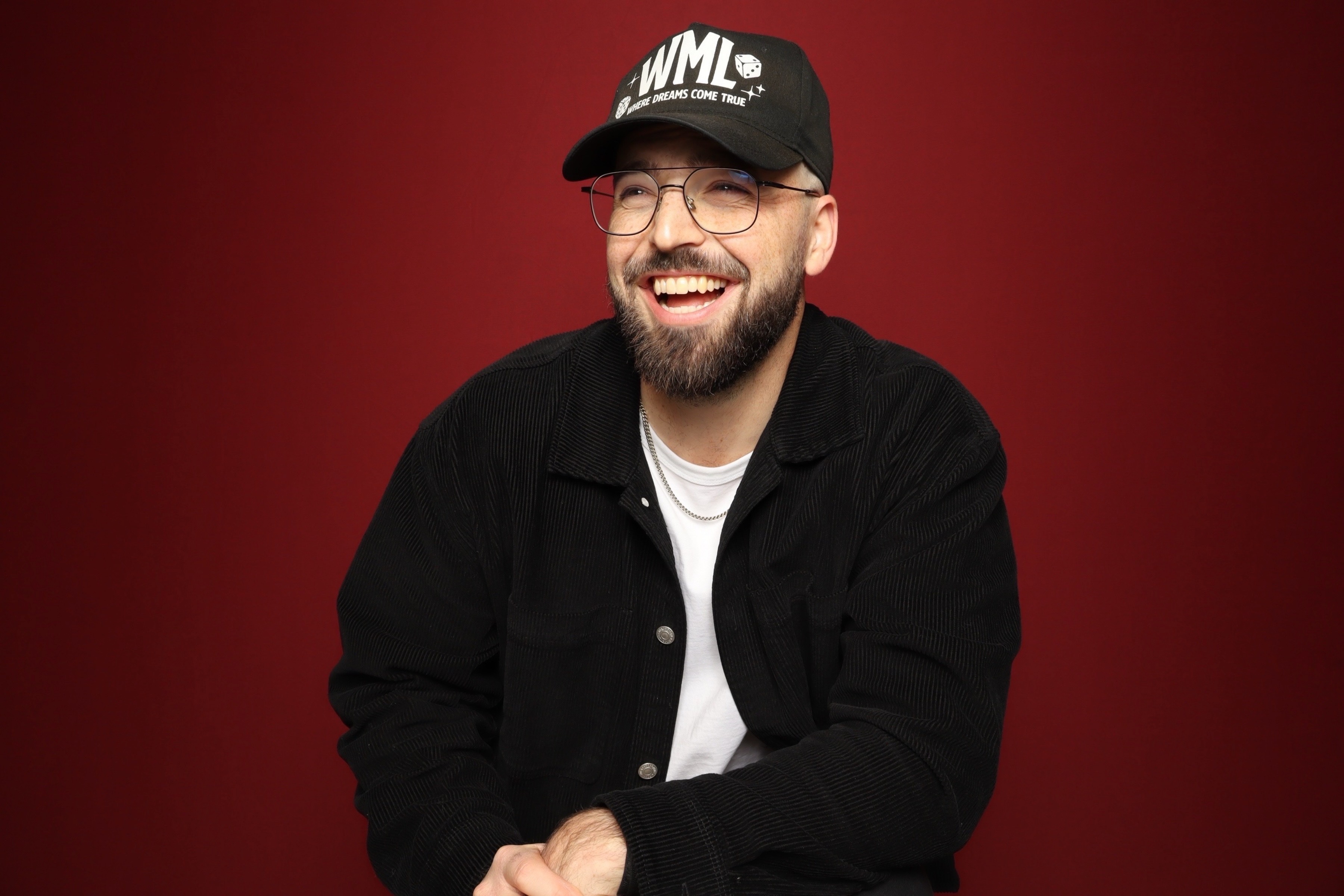 Portrait photo booth photo of a smiling bearded man wearing glasses, a black 'Creative Department' cap, black jacket, and a gold chain necklace in front of a green textured background.
