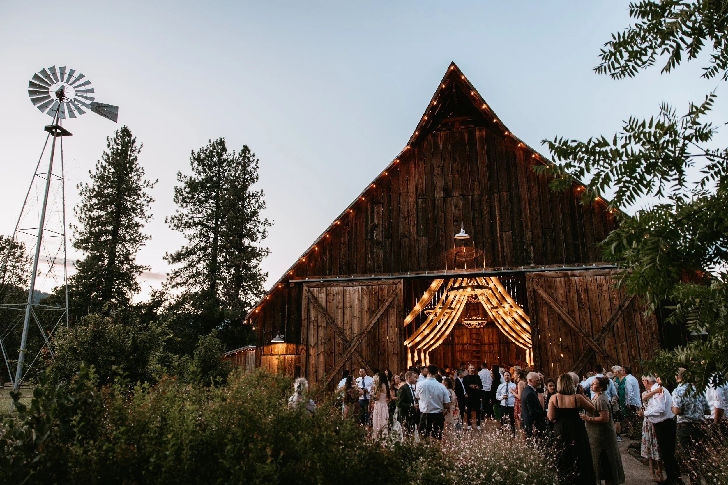 Guests socializing outside and inside a large rustic wooden barn decorated with draped lights at dusk, with a windmill and tall trees nearby.