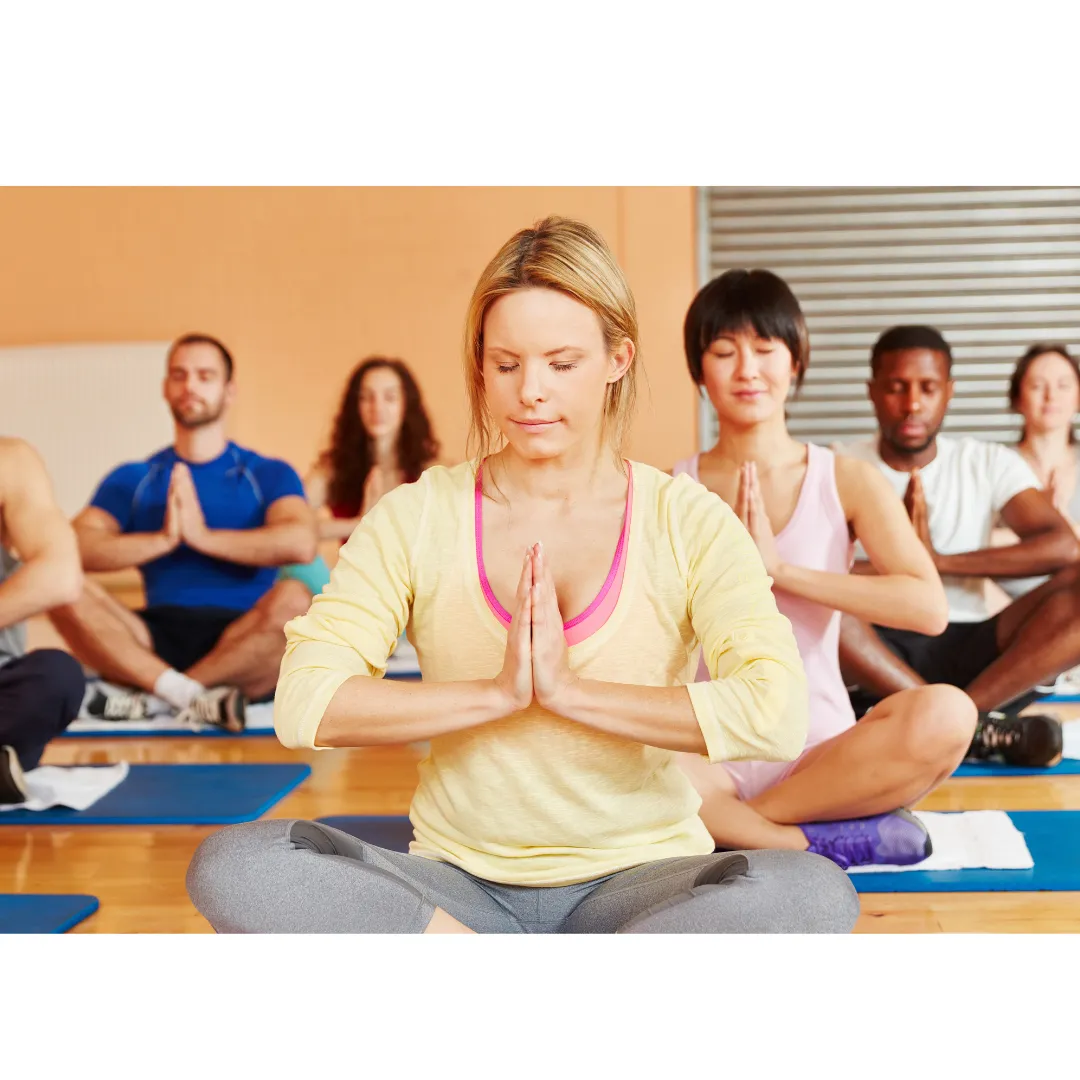 several yoga students sitting on yoga mats with their eyes closed and their hands in prayer position in front of their hearts