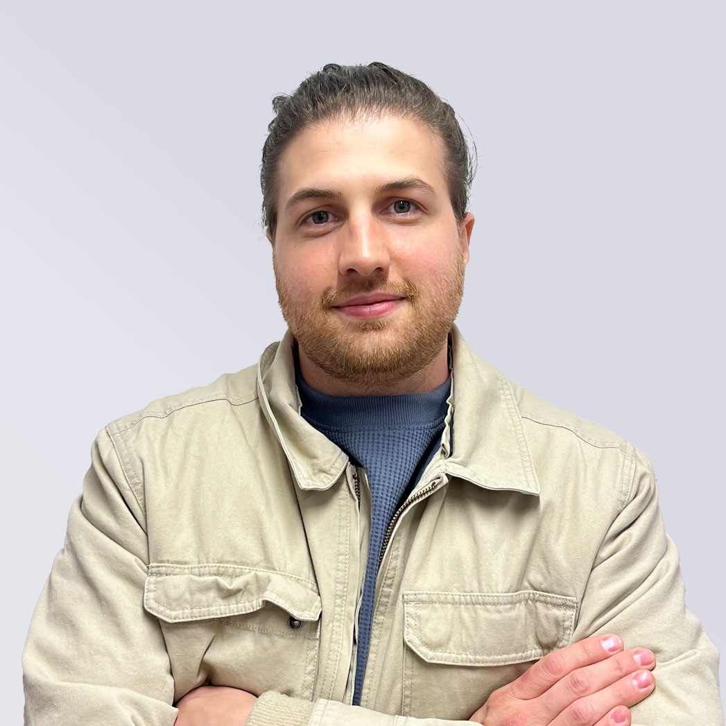 Smiling man with beard and tied-back hair wearing a beige jacket and navy shirt, arms crossed.