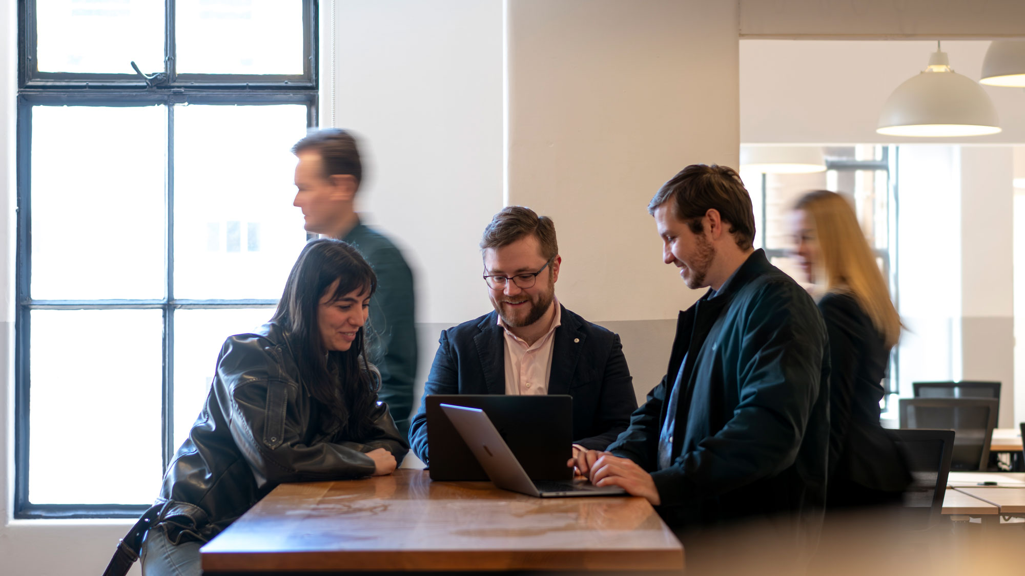 Three people sitting at a table, two using laptops, engaged in discussion inside an office.