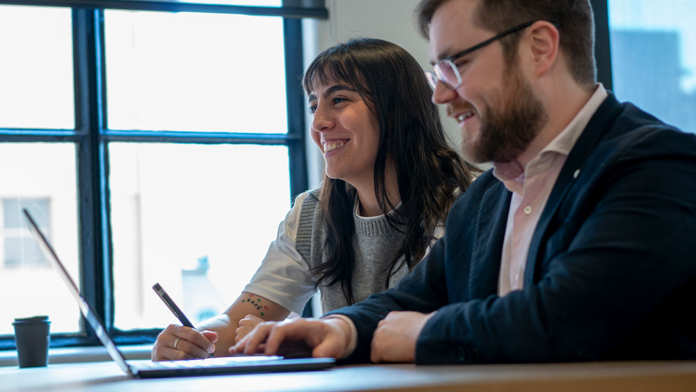 Smiling man and woman working together on a laptop at a table in a bright office.