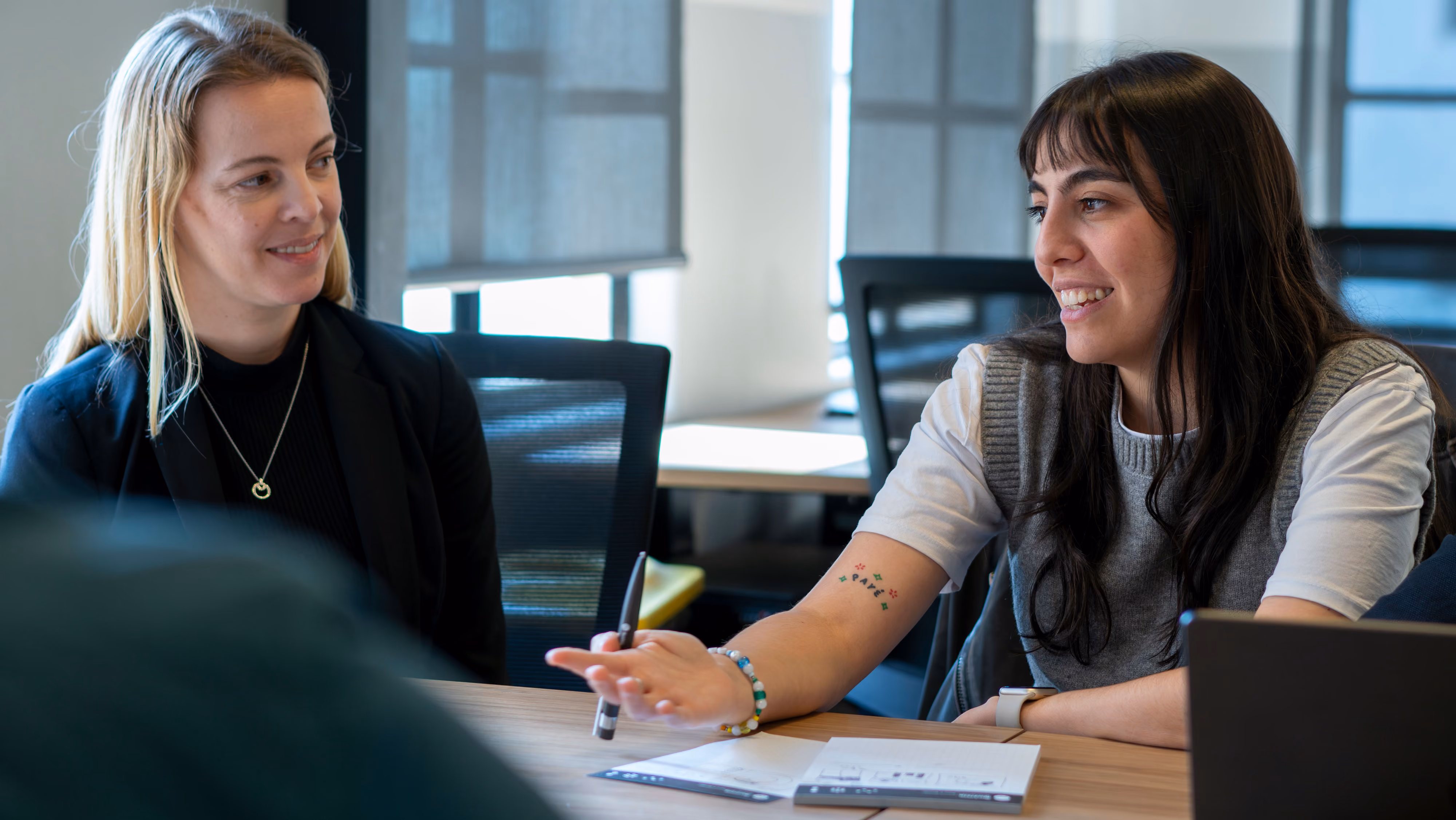 Two women seated at a table in a meeting room, one speaking and gesturing while the other listens.