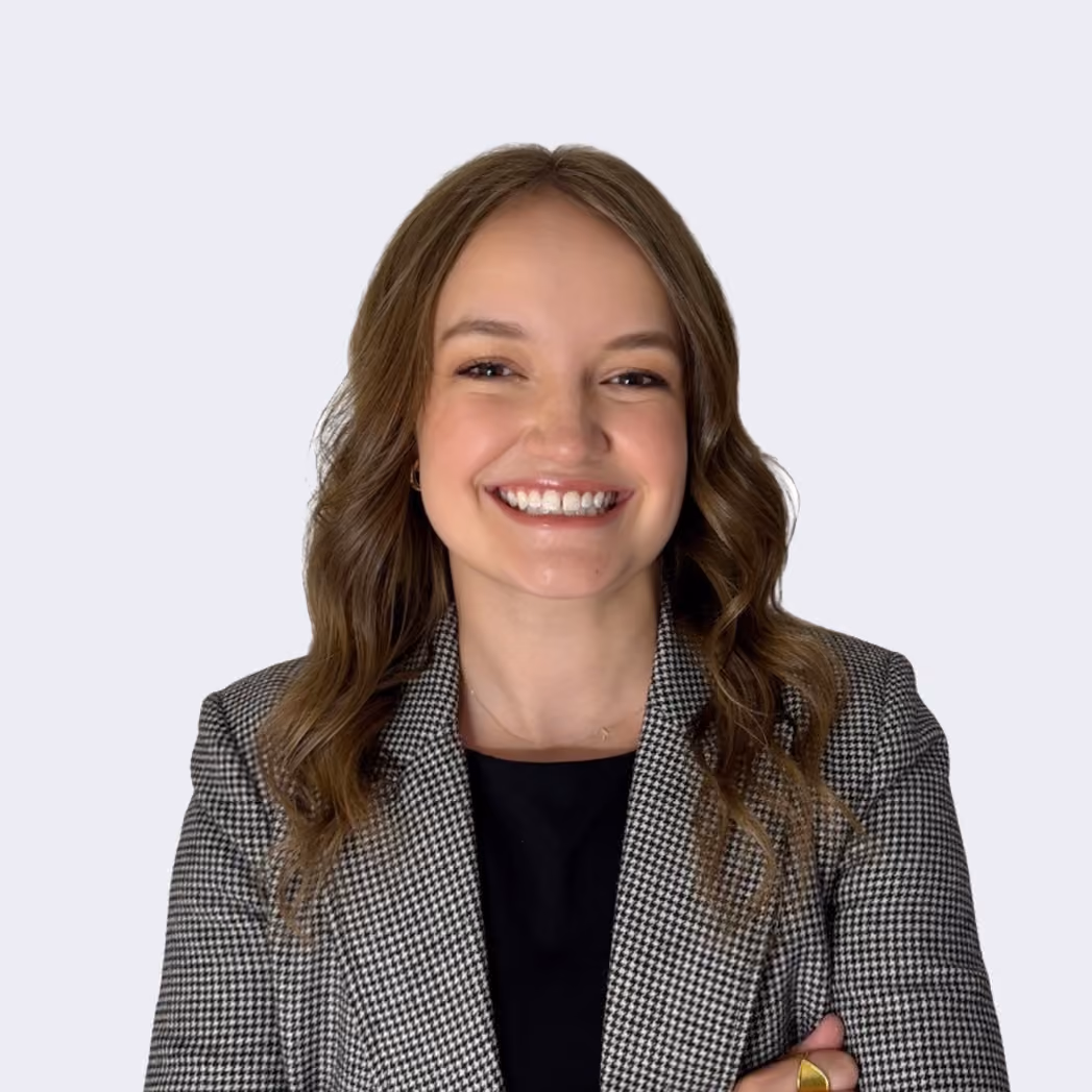 Smiling young woman with light brown wavy hair wearing a black top and a grey checkered blazer, standing against a plain light background.