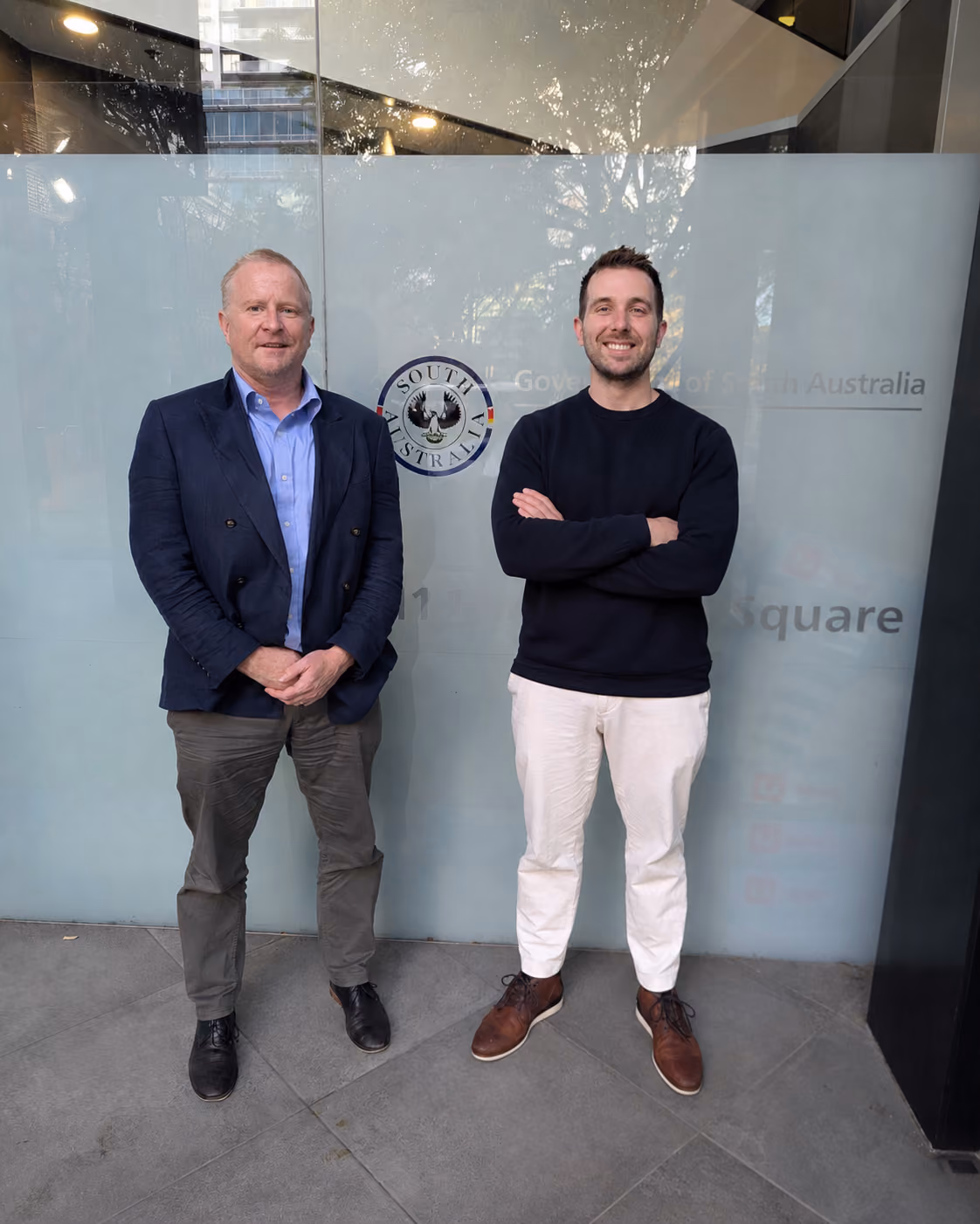 Two men standing side by side in front of a frosted glass wall with the South Australia government logo, one man wears a navy blazer with a light blue shirt and grey pants, the other wears a black sweater with white pants and brown shoes.