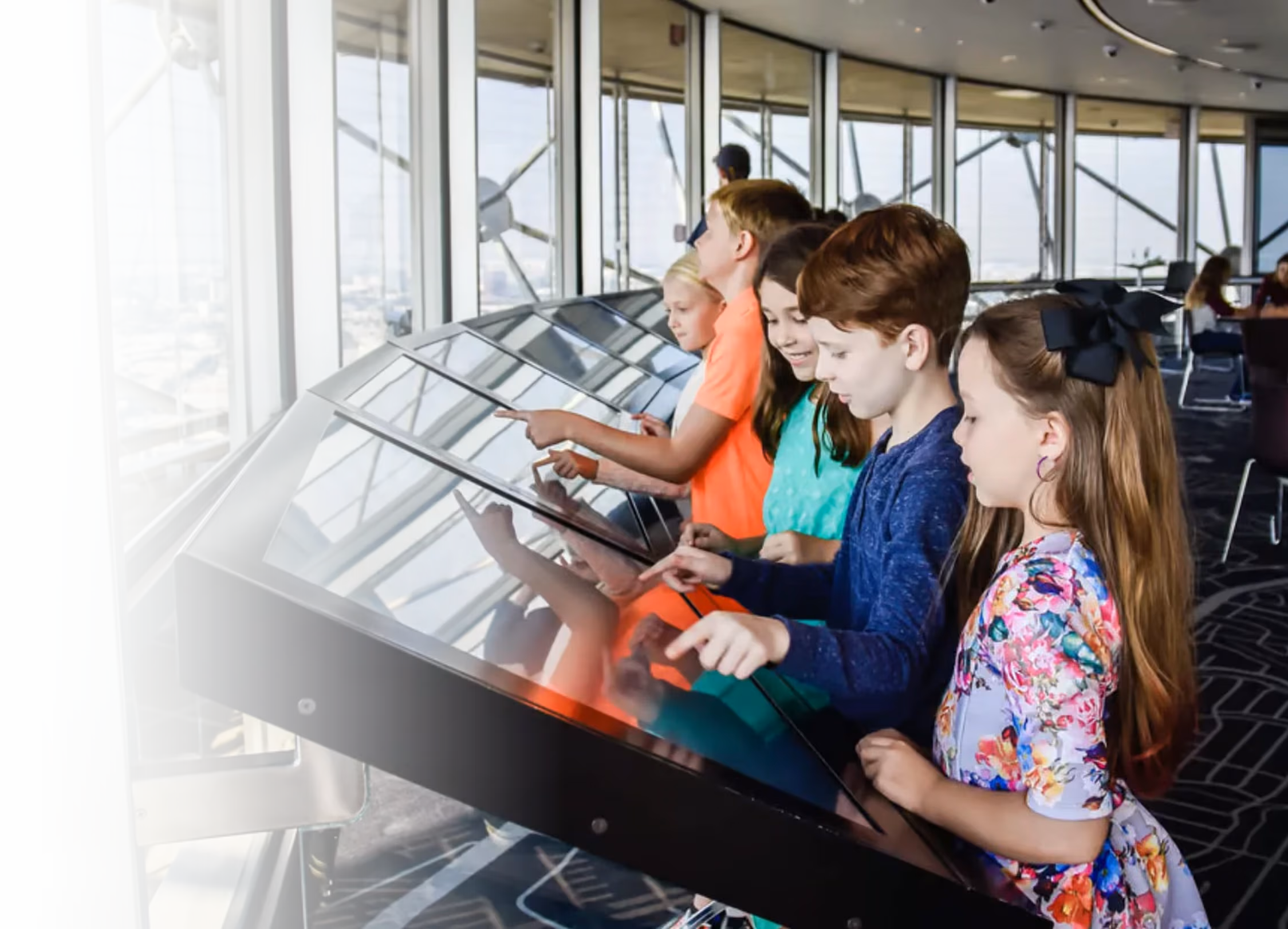 Children interact with touchscreen displays at an observation deck with panoramic city views
