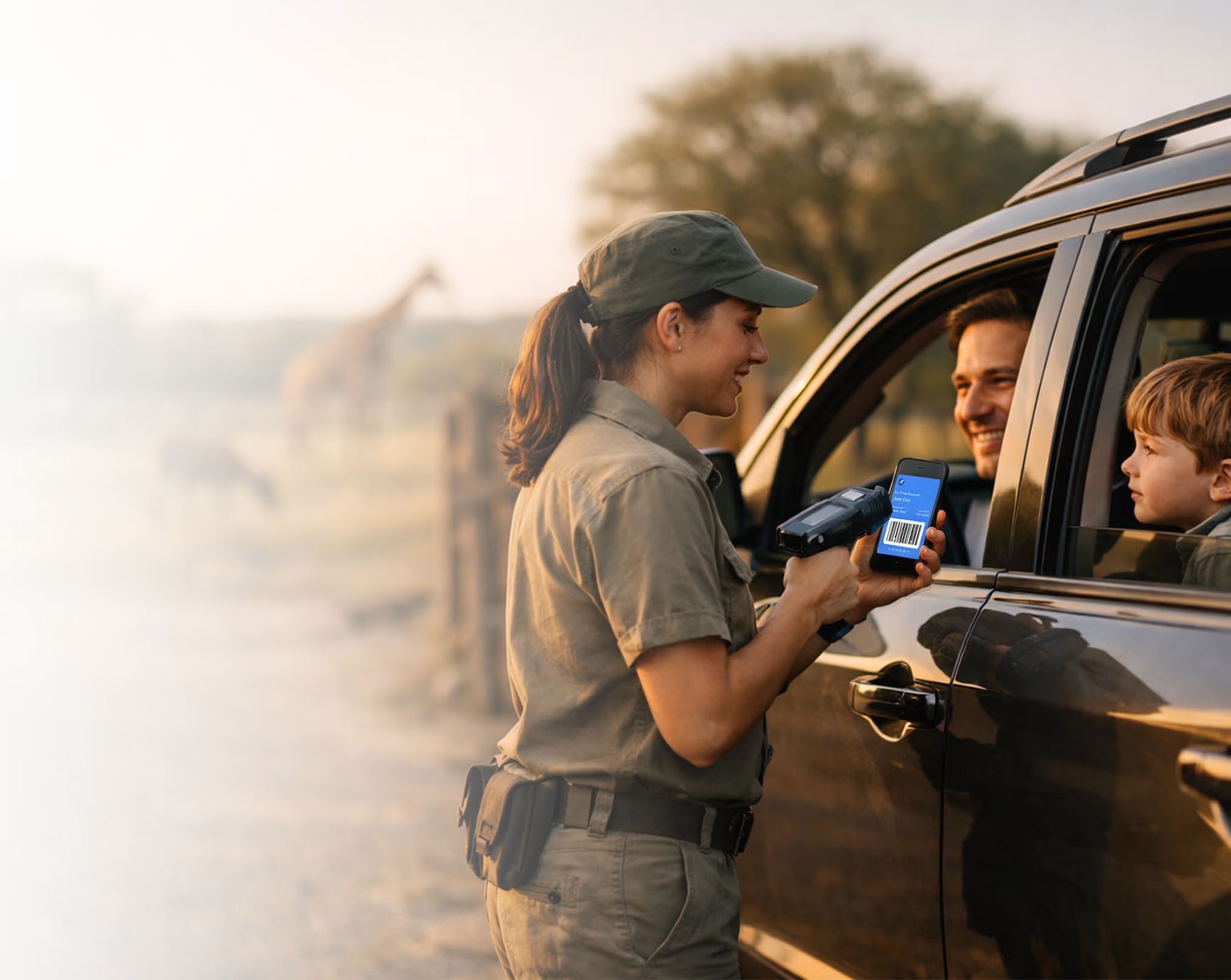 Wildlife park staff scanning a family's mobile ticket at a drive-through safari entrance with giraffes nearby.