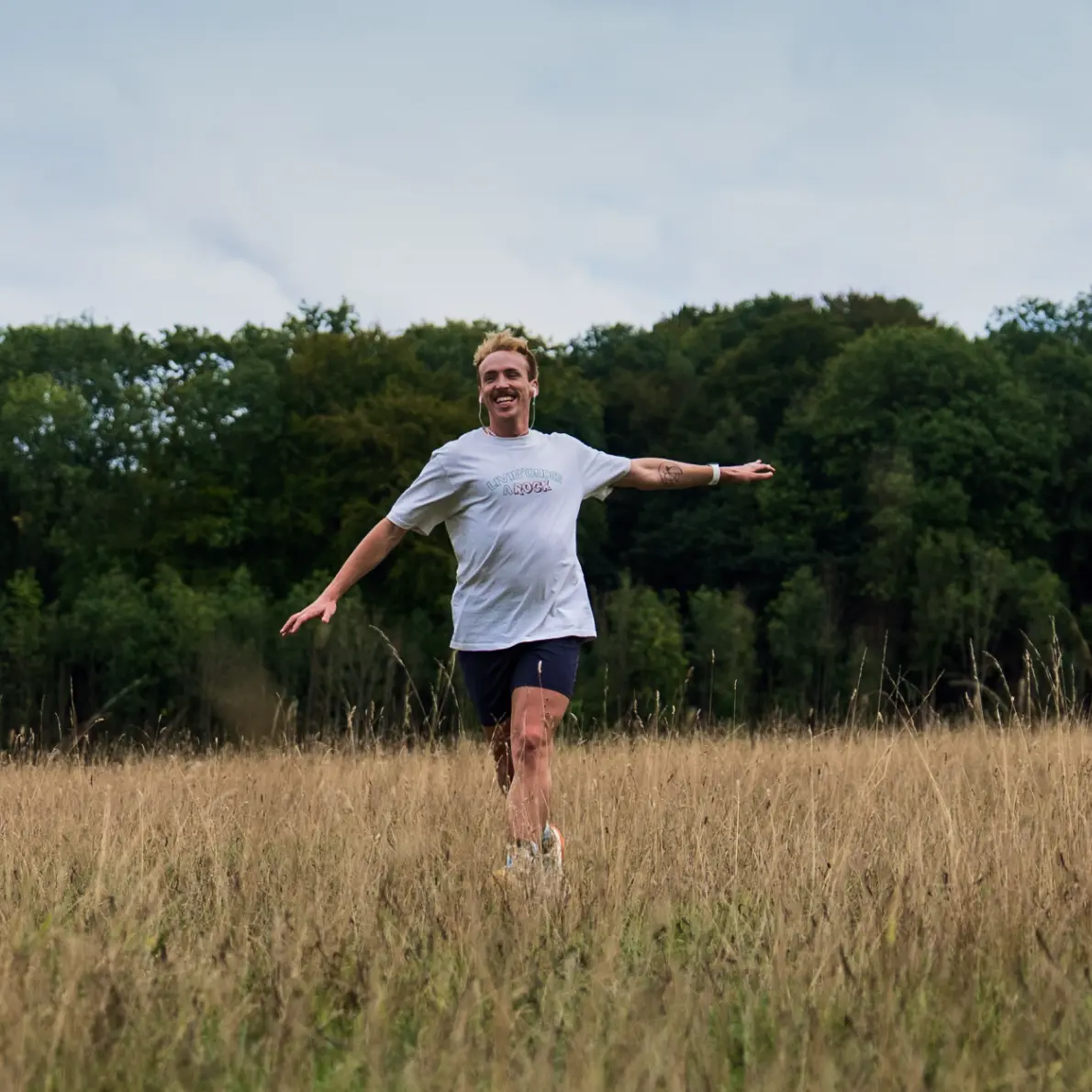 A person with a mustache happily running with arms outstretched through a tall grass field, wearing a white t-shirt, dark shorts, and earphones, with a forest in the background under a cloudy sky.