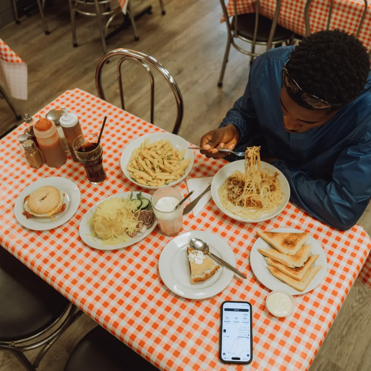 Person in a dark jacket eating spaghetti at a table covered with an orange and white checkered tablecloth, surrounded by plates of pasta, a sandwich, toast, a dessert slice, condiments, and drinks, with a smartphone showing a graph on the table.