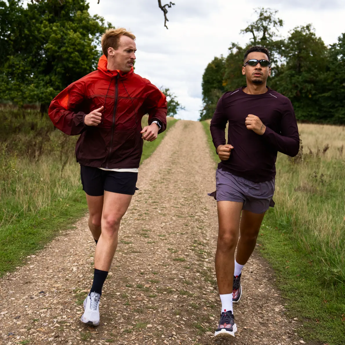 Two men running on a gravel path surrounded by grass and trees, one wearing a red jacket and the other wearing a dark long-sleeve shirt with sunglasses.