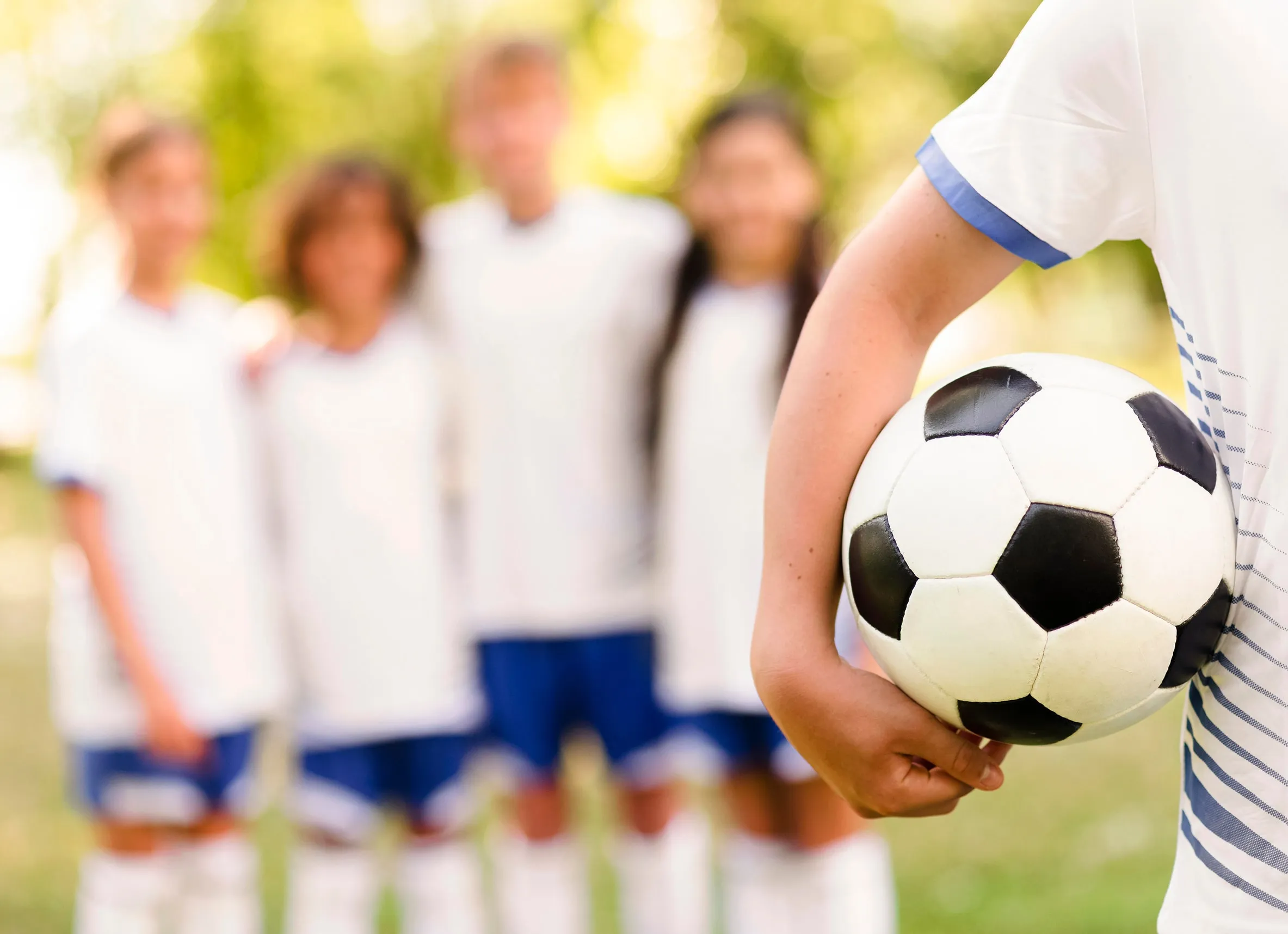 Close-up of a person holding a black and white soccer ball with a blurry background of four kids in white soccer uniforms standing side by side outdoors.