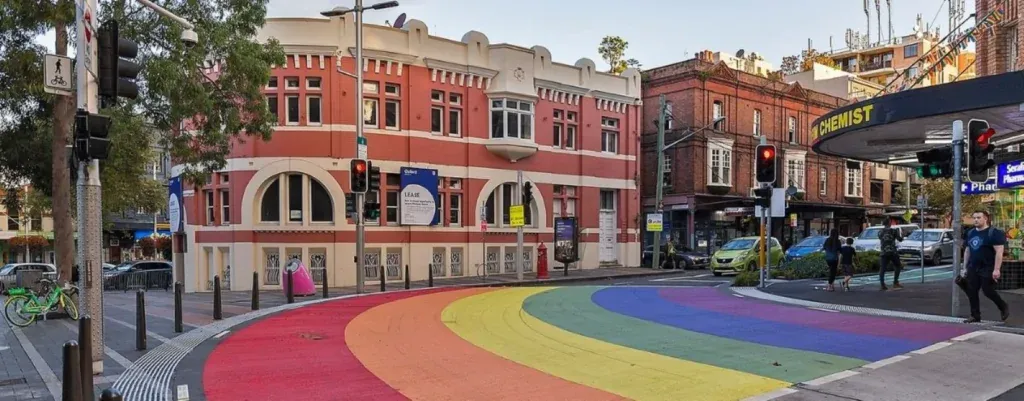 A rainbow-colored crosswalk spans a city street in Surry Hills beside red brick buildings.