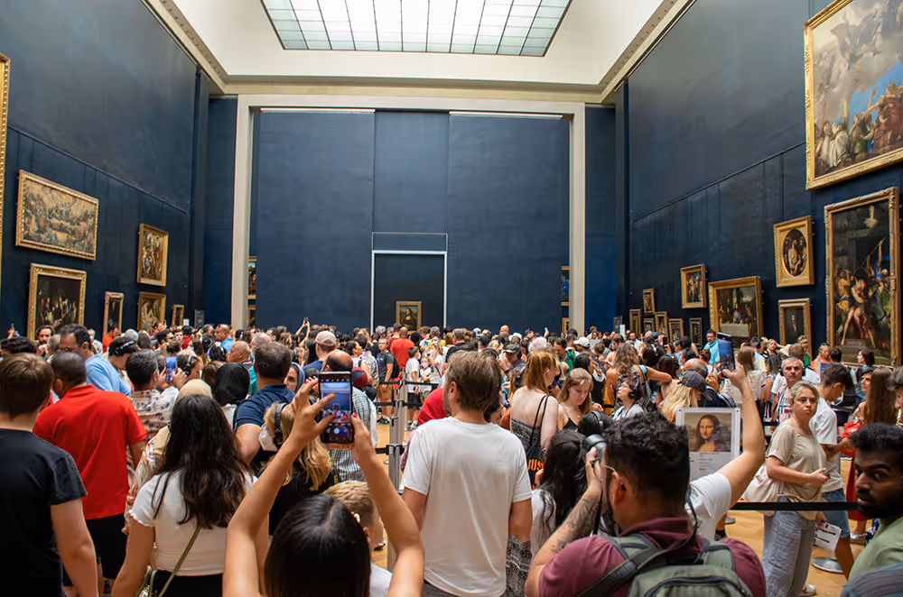 Crowded gallery room in a museum with many people looking at the Mona Lisa painting on the far wall.