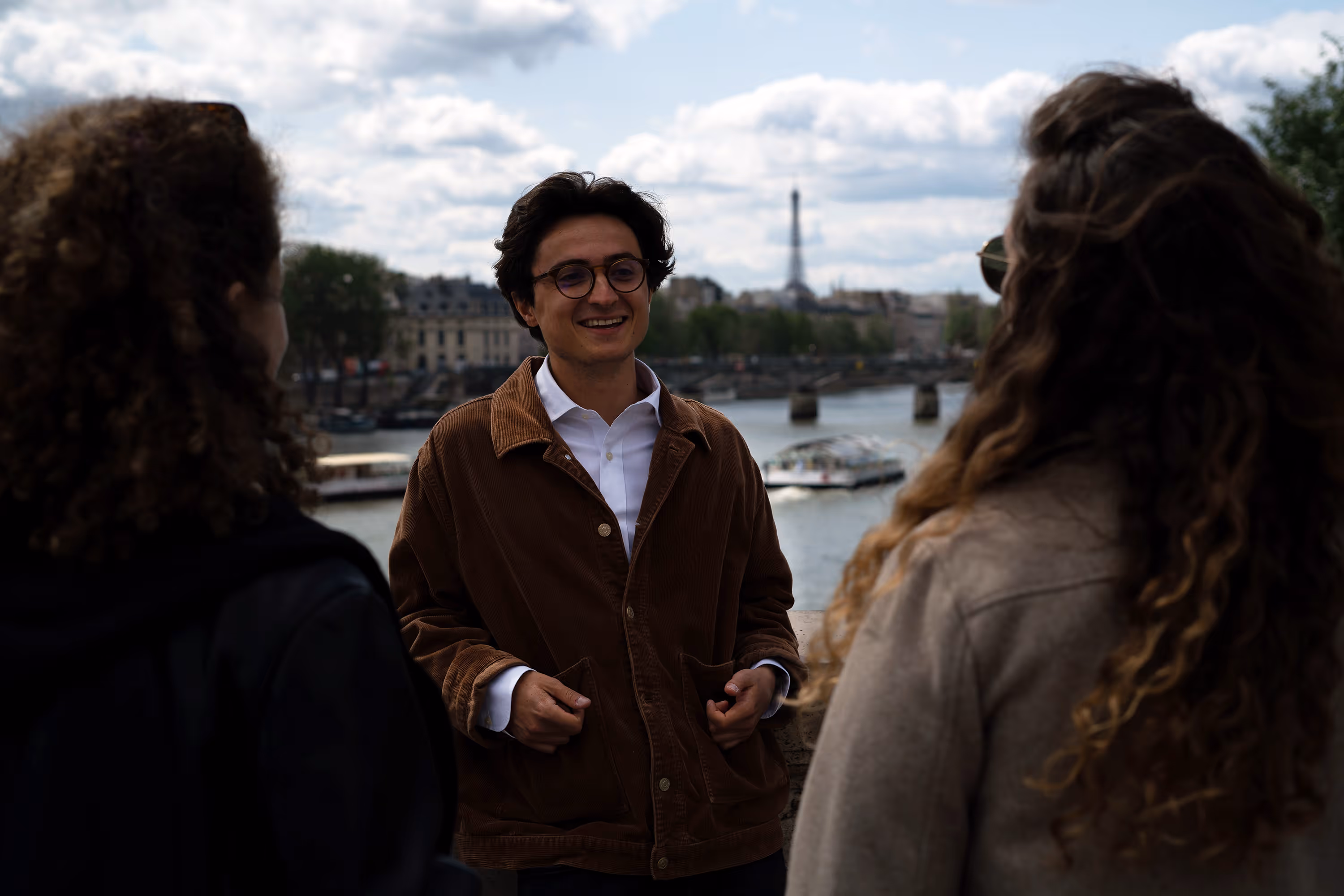 Smiling man wearing glasses and brown jacket talking to two women with curly hair near a river with the Eiffel Tower in the background.