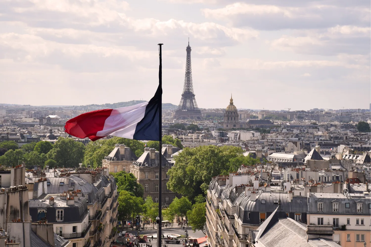 French flag at half-mast with the Eiffel Tower and Paris skyline in the background under a cloudy sky.