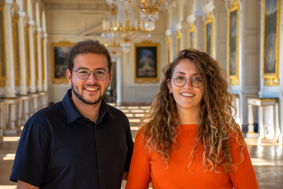 Smiling man and woman with glasses standing in a bright, ornate hallway with chandeliers.
