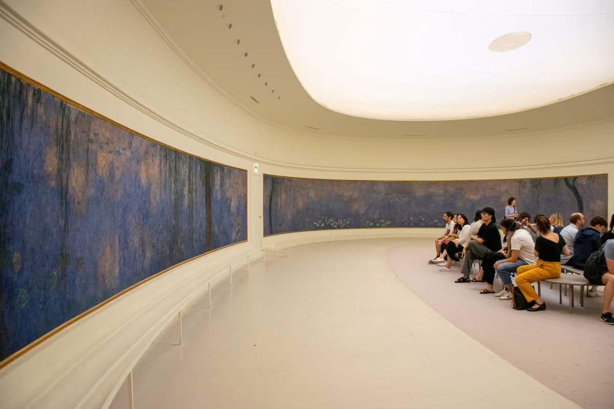 People seated on benches in a curved gallery viewing large blue-toned paintings.