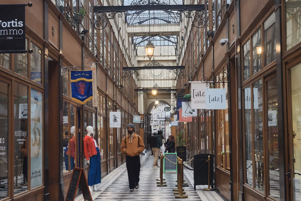 Man walking in a covered shopping arcade with glass ceiling and shops on both sides.
