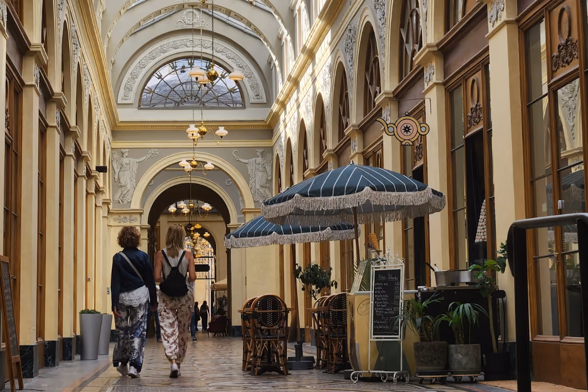 Two people walking in a covered arcade with outdoor café seating and striped umbrellas.