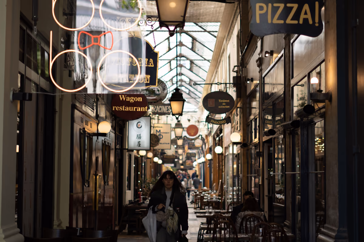 Narrow covered alley lined with restaurant signs and tables, with a woman walking toward the camera.