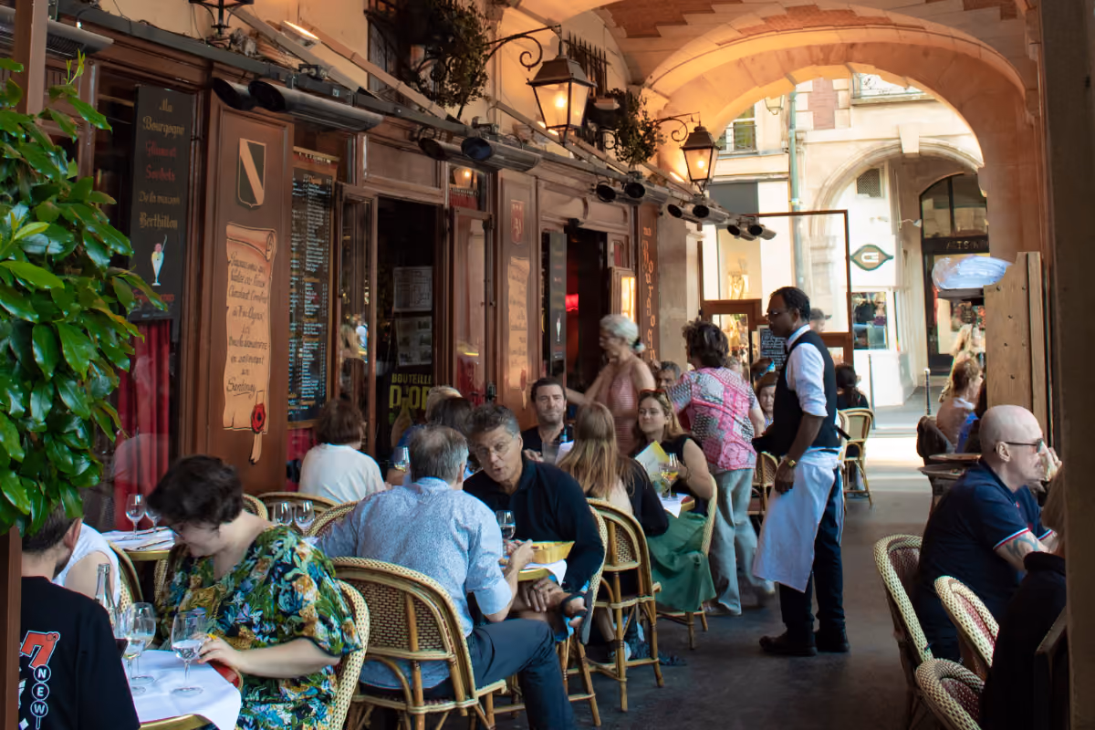 People dining and a waiter serving at an outdoor cafe under an arched walkway with plants and signs.