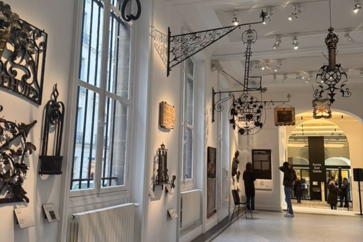 Museum gallery with ironwork exhibits on walls and ceiling, visitors observing displays near windows.