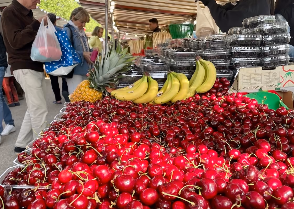 Large pile of fresh cherries with bananas and a pineapple on a market stall with shoppers in background.