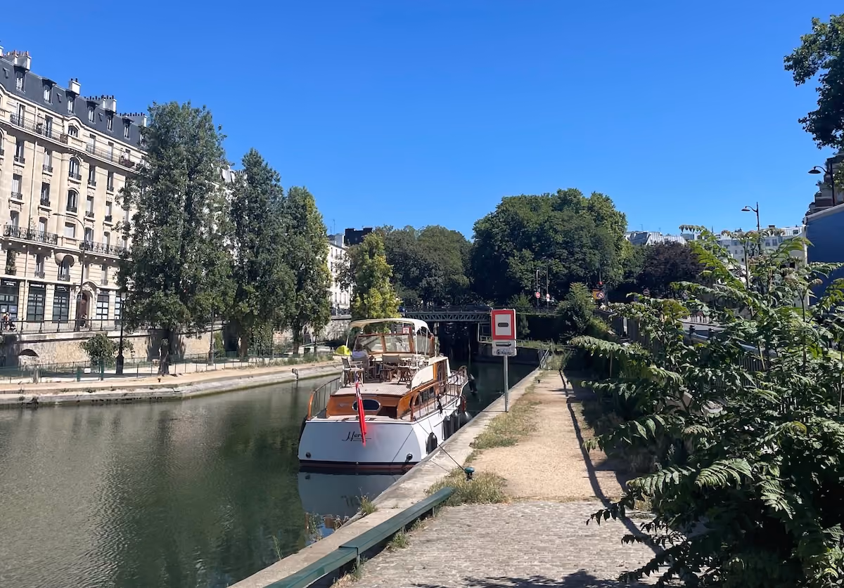 Boat docked on a canal with trees and classic buildings under a clear blue sky.