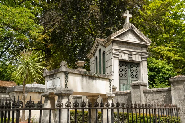 Mausoleo de piedra con una cruz en la parte superior y un sarcófago de piedra elevado rodeado por una verja de hierro negro, situado entre árboles verdes en un cementerio.