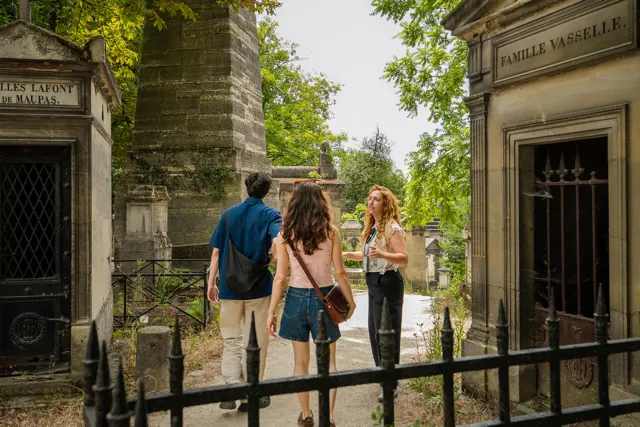 Tres jóvenes adultos caminando por un sendero del cementerio entre mausoleos con puertas de hierro, rodeados de vegetación bajo un cielo parcialmente nublado.