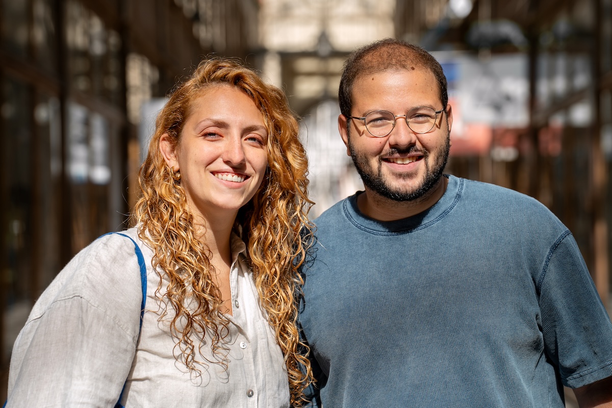 Mujer sonriente con cabello rubio rizado y camisa gris claro, de pie junto a un hombre sonriente con gafas y cabello corto, vestido con una camisa azul, en un mercado interior o pasaje iluminado por el sol.