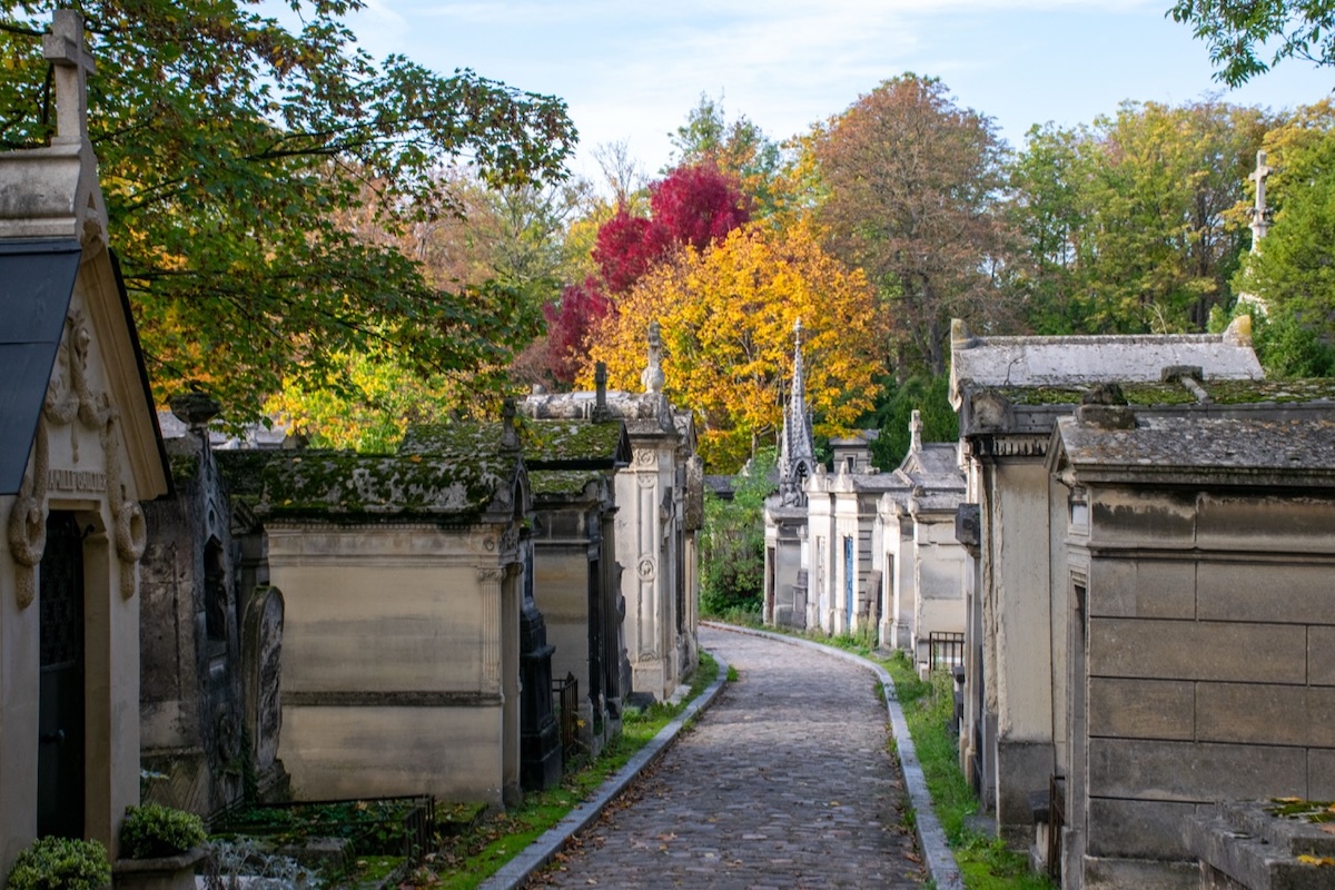 Un camino de adoquines que serpentea por un cementerio histórico bordeado de antiguos mausoleos y tumbas de piedra, con árboles de colores otoñales al fondo.