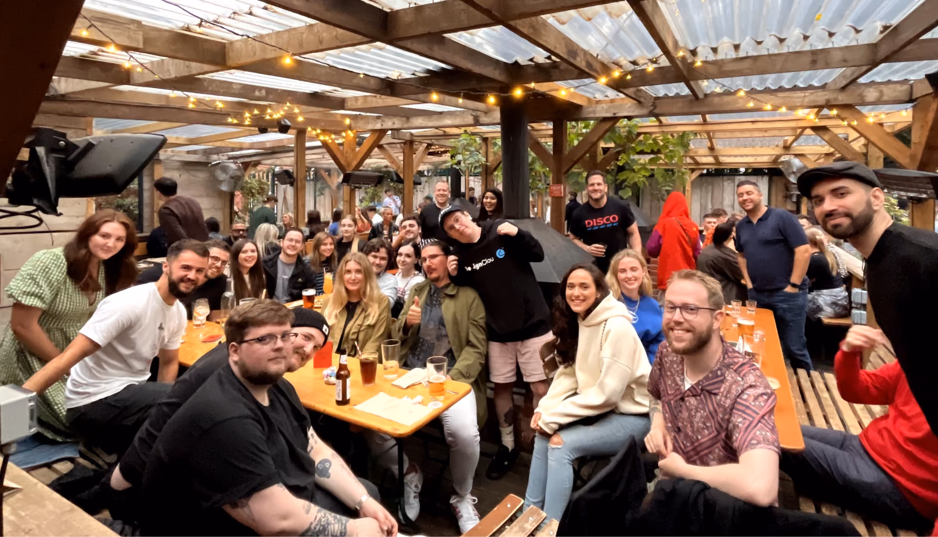 Group of people smiling and sitting at tables with drinks under a covered outdoor patio with string lights.