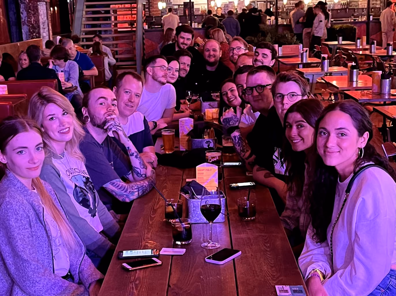 Group of people sitting at a long table in a bar, smiling at the camera with drinks on the table.