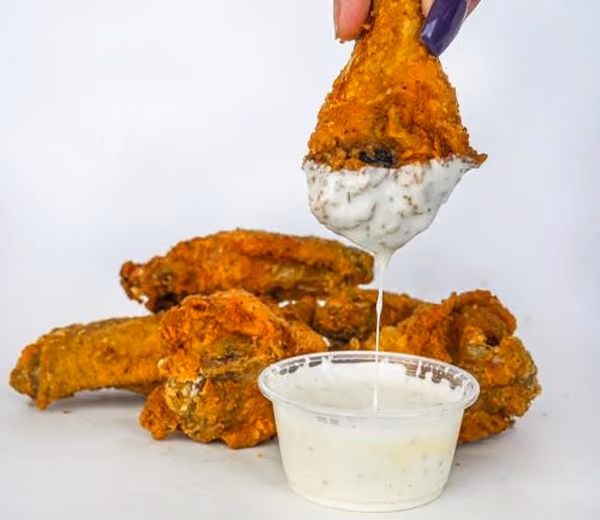 A hand with purple nails dips a crispy, golden-brown chicken wing into white ranch sauce, with more wings in the background, creating a tempting and savory visual.