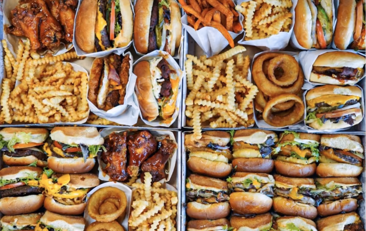 An array of fast food items, including burgers, wings, fries, sweet potato fries, and onion rings, neatly arranged in boxes. The scene conveys abundance and indulgence.