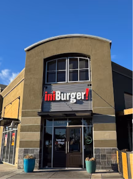 Exterior of iniBurger restaurant with glass entrance, two blue planters, under clear blue sky.