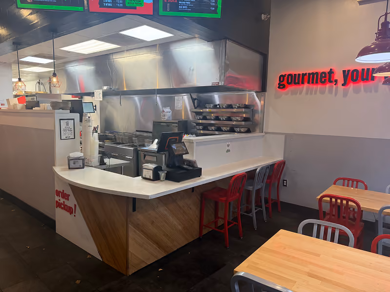 Interior view of a restaurant kitchen area with a worker cooking and tables with chairs in the dining area.