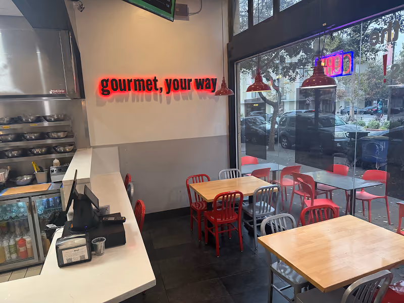 Interior of a modern café with a wooden counter, digital registers, and seating near large windows showing parked cars outside.