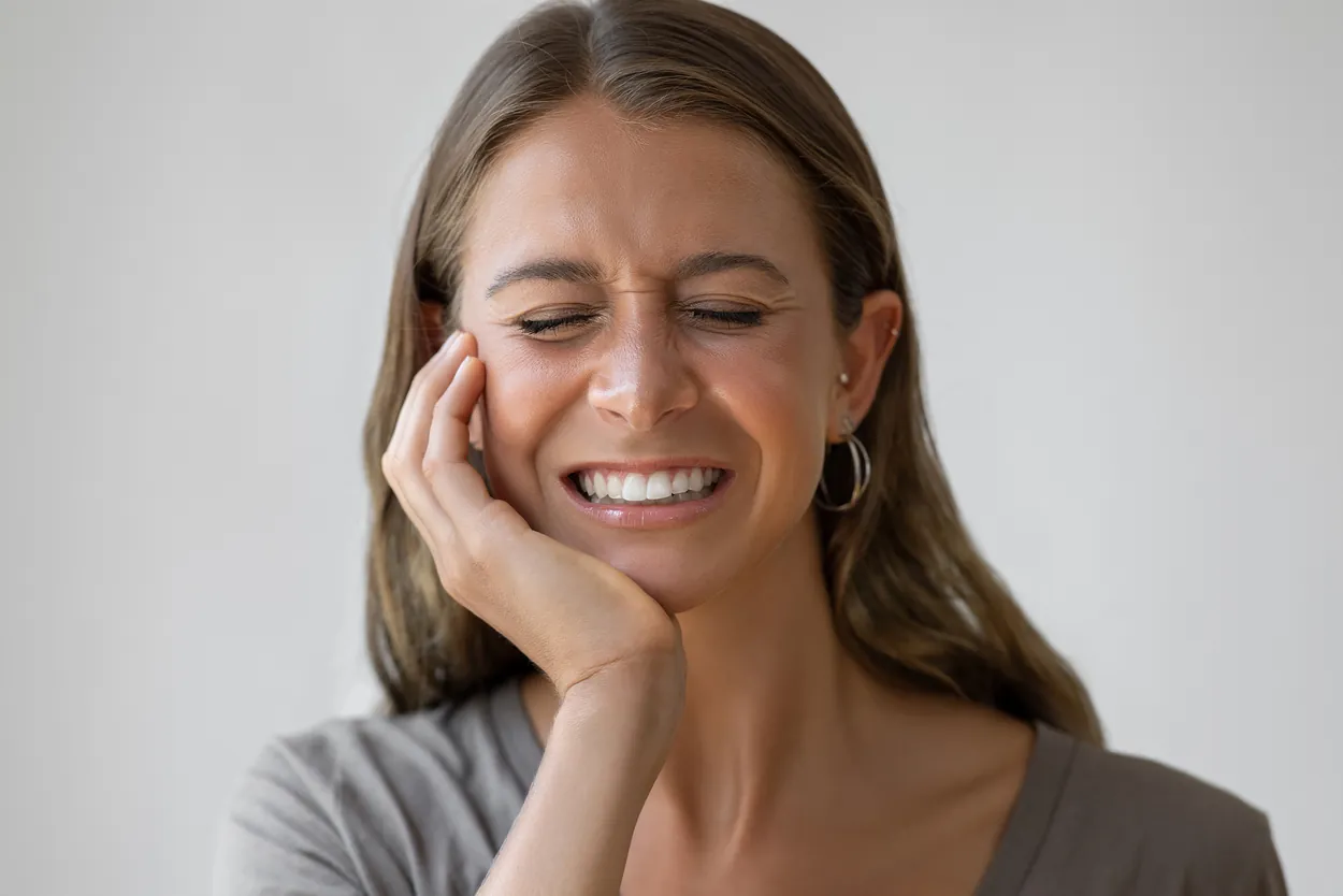 Woman in soft lighting grimacing and holding her jaw, conveying toothache or dental pain against a neutral grey background.