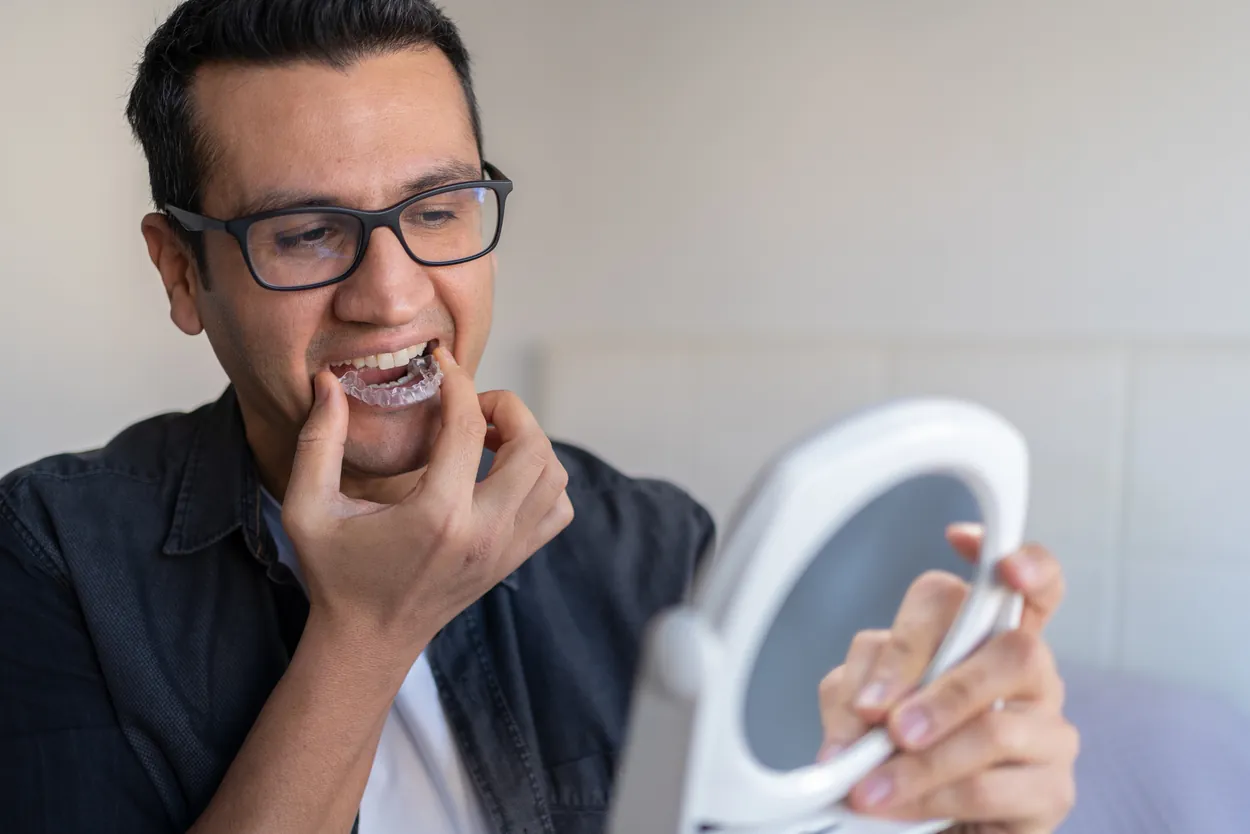 Man with glasses using a handheld mirror to carefully insert a clear orthodontic aligner, captured in bright, soft natural indoor lighting.
