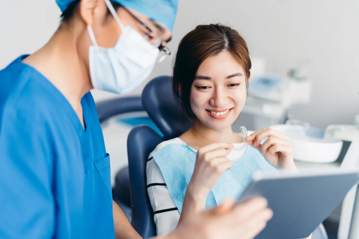 Dentist in blue scrubs and surgical mask reviews digital treatment plan on a tablet with a smiling patient holding clear aligners in a bright clinic.