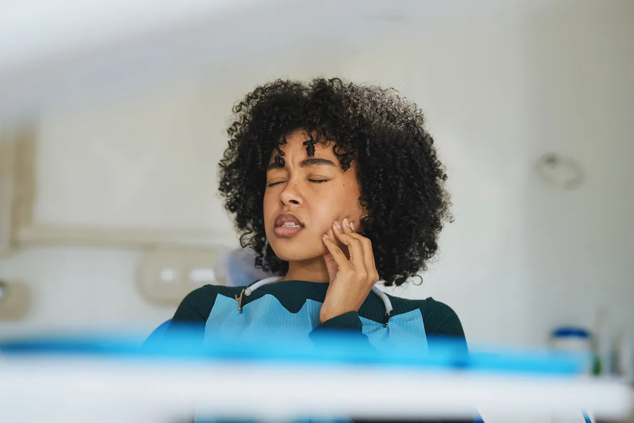 Young woman with curly hair wearing a dental bib and experiencing tooth pain in a brightly lit, clinical dental office setting.