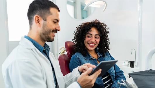 Smiling dentist in a white coat shows a digital tablet to a patient in a bright, modern dental office, emphasizing personalized clinical consultation.